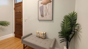A hallway with a wooden door, a grey bench with a patterned cushion, a framed artwork above the bench, and a large green plant to the right. - Cozy 3BR Gem in North Center (Chicago)
