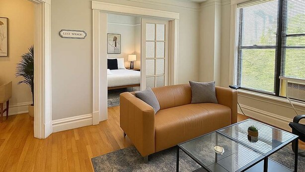 A room with a brown leather sofa, a glass coffee table, and a bed in the background. - Cozy 3BR Gem in North Center (Chicago)