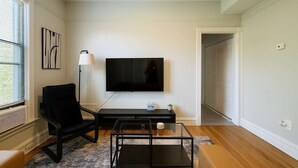 A living room with a flat-screen TV mounted on the wall, a black chair, a glass coffee table, and a small side table with a lamp and a plant. - Cozy 3BR Gem in North Center (Chicago)