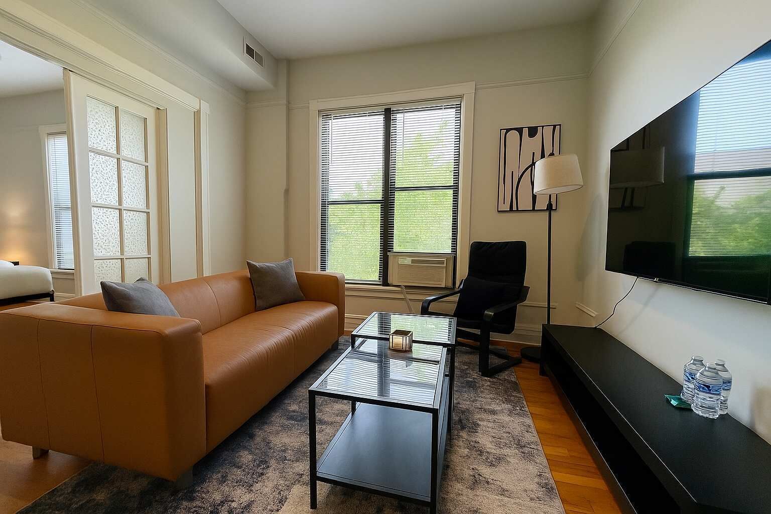 A modern living room with a leather sofa, a glass coffee table, a black office chair, a flat-screen TV, and a large window with blinds.