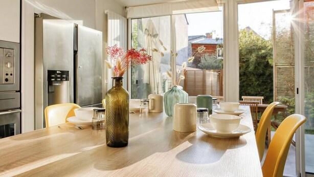 Eine moderne Küche mit Edelstahlgeräten, einem Holzespeisetisch mit gelben Stühlen und Blick auf den Garten durch große Fenster.