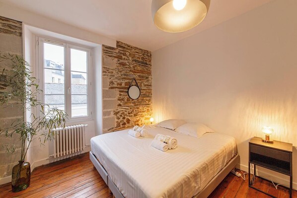 A bedroom with a stone wall, a large window, a bedside table with a lamp, and a bed with rolled towels.