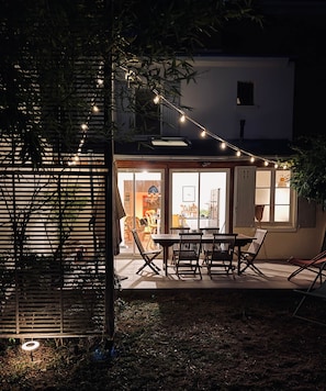 A patio area with a table and chairs, string lights, and a view of the interior through glass doors.