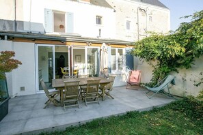 Un patio con una mesa y sillas de madera, un vidrio que da a una habitación y un pequeño jardín con una silla y una hamaca.