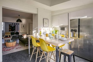 Una cocina moderna con un refrigerador de acero inoxidable, un microondas y un dispensador de agua. Hay una zona de comedor con sillas amarillas y una mesa de madera.