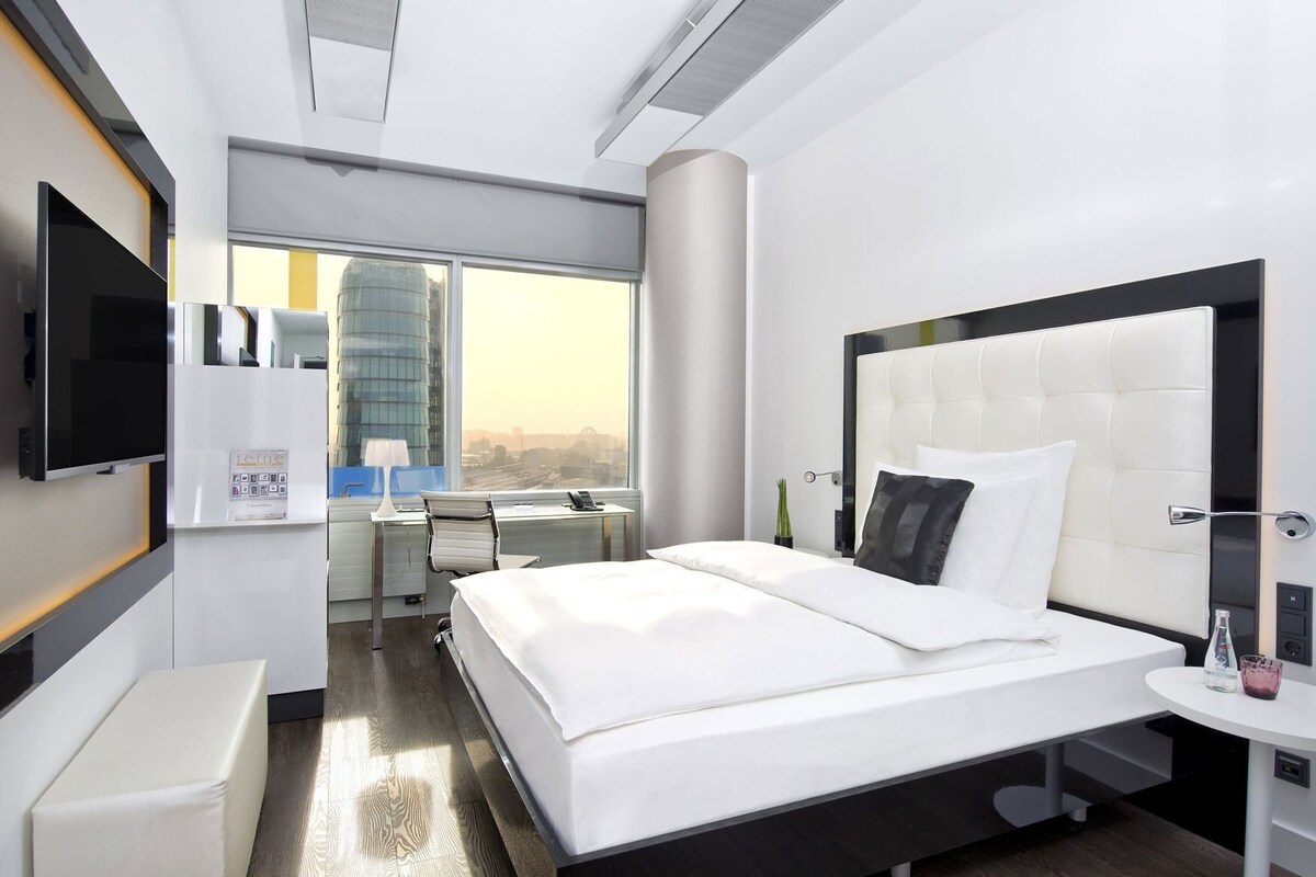 A modern hotel room with a large bed, a desk, a chair, a TV, and a window with a city view.