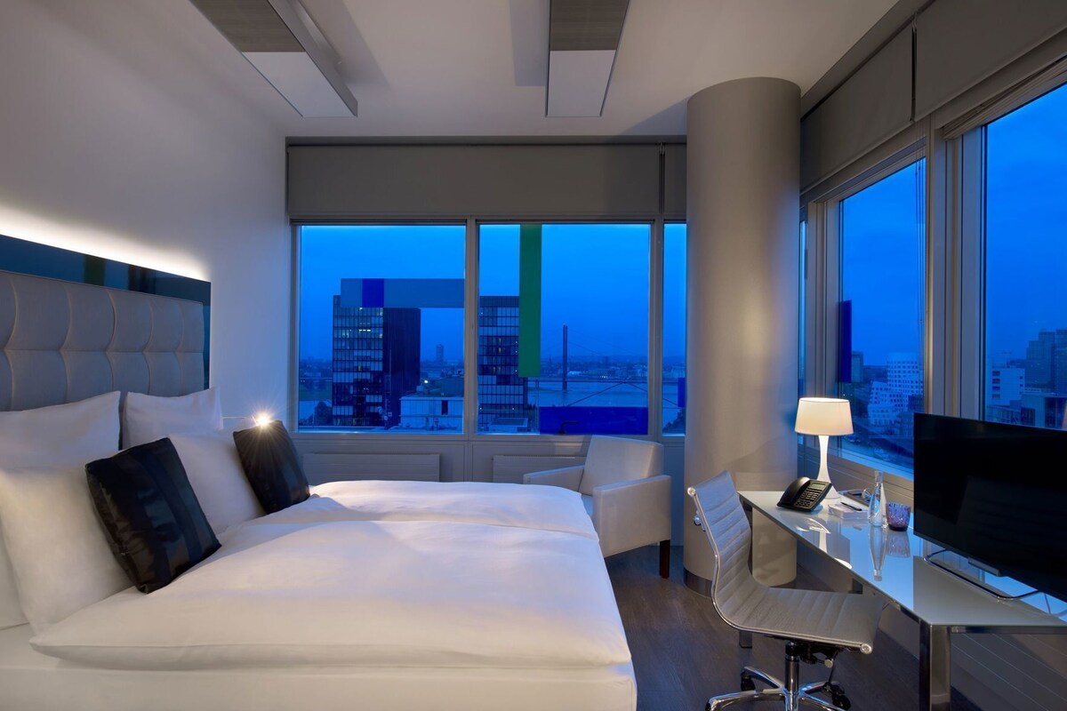 A hotel room with a bed, a desk, a chair, a TV, and a large window with a city view.