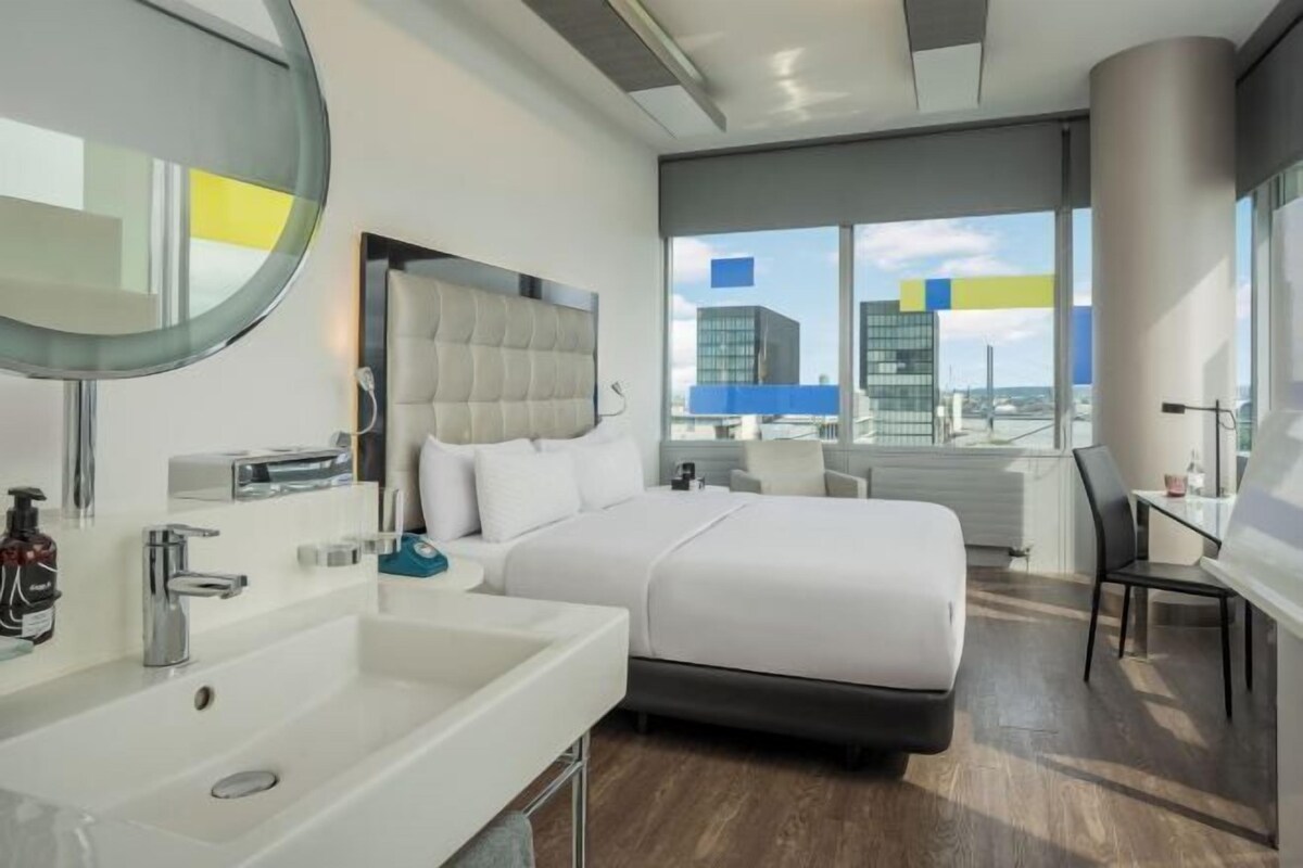 Une chambre d’hôtel moderne avec un grand lit, un miroir rond, un lavabo blanc et une vue sur le paysage urbain.