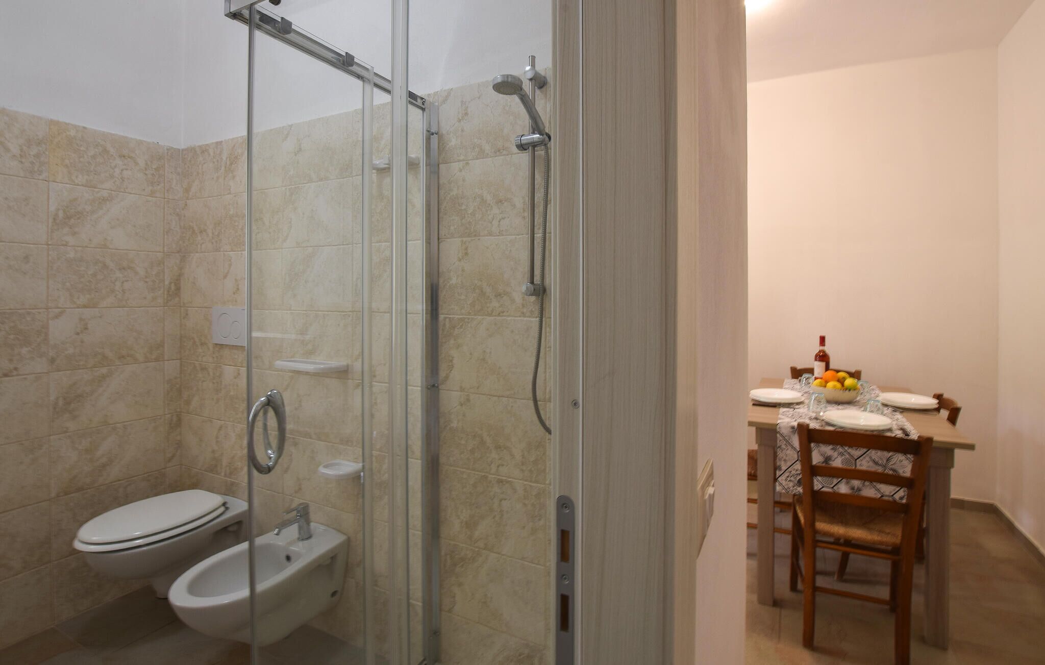 A bathroom with a glass-enclosed shower, a toilet, and a bidet. There is a dining area with a table set for two, a bowl of fruit, and a bottle of wine.