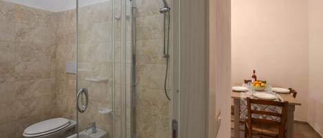 A bathroom with a glass-enclosed shower, a toilet, and a bidet. There is a dining area with a table set for two, a bowl of fruit, and a bottle of wine.
