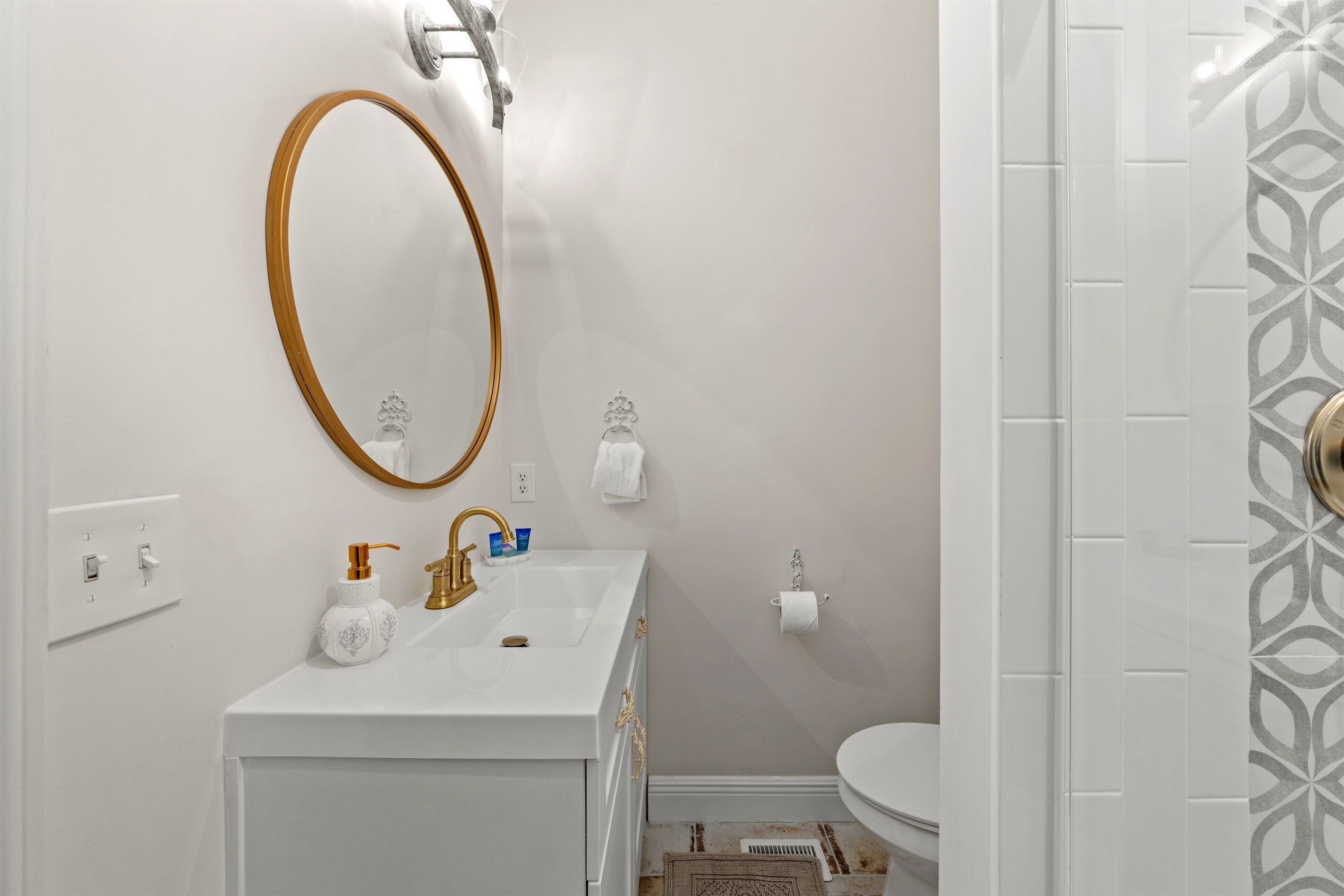 A bathroom with a white sink, gold faucet, and a large oval mirror. There is a toilet with a white towel and a paper towel holder.