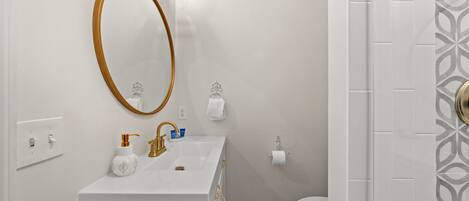 A bathroom with a white sink, gold faucet, and a large oval mirror. There is a toilet with a white towel and a paper towel holder.