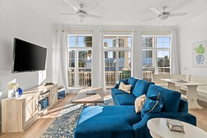 Uma sala de estar ampla com um sofá azul grande, uma TV de tela plana e um espaço de jantar com mesa e cadeiras brancas.