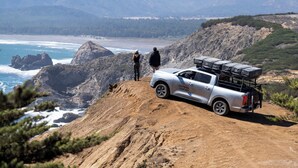 Un camionnette argentée est garée sur un chemin de terre, avec une falaise rocheuse et l’océan en arrière-plan.