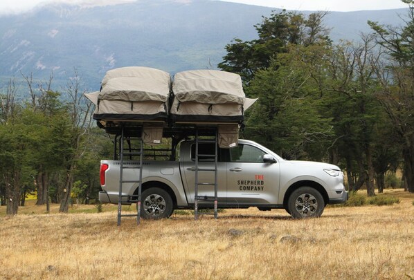 A silver pickup truck with a bed covered in a tarp, parked in a grassy field with trees and mountains in the background. - The Shepherd Company (Coyhaique)