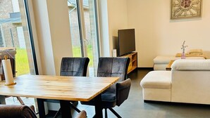 Ein modernes Wohnzimmer mit Essbereich, ausgestattet mit einem Holztisch und Stühlen, einem weißen Sofa und einem Flachbildfernseher auf einem Holzgestell.