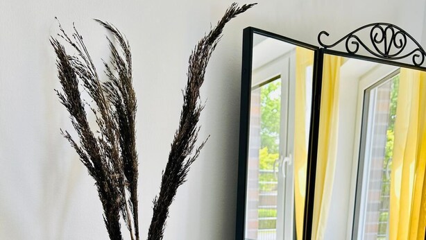 Ein Zimmer mit einem Spiegel, der ein Fenster mit gelben Vorhängen, eine blaue Vase mit getrocknetem Pampasgras, eine gelbe Vase mit einer weißen Blume und eine Duftkerze reflektiert.