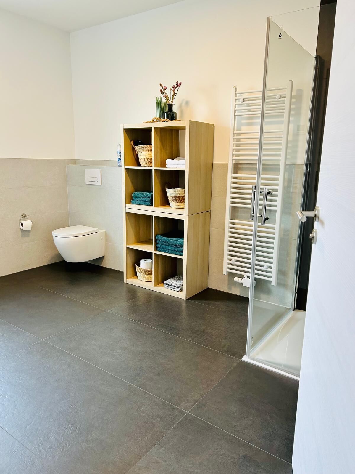 A modern bathroom with a toilet, a wooden shelf unit with baskets, and a glass shower enclosure.