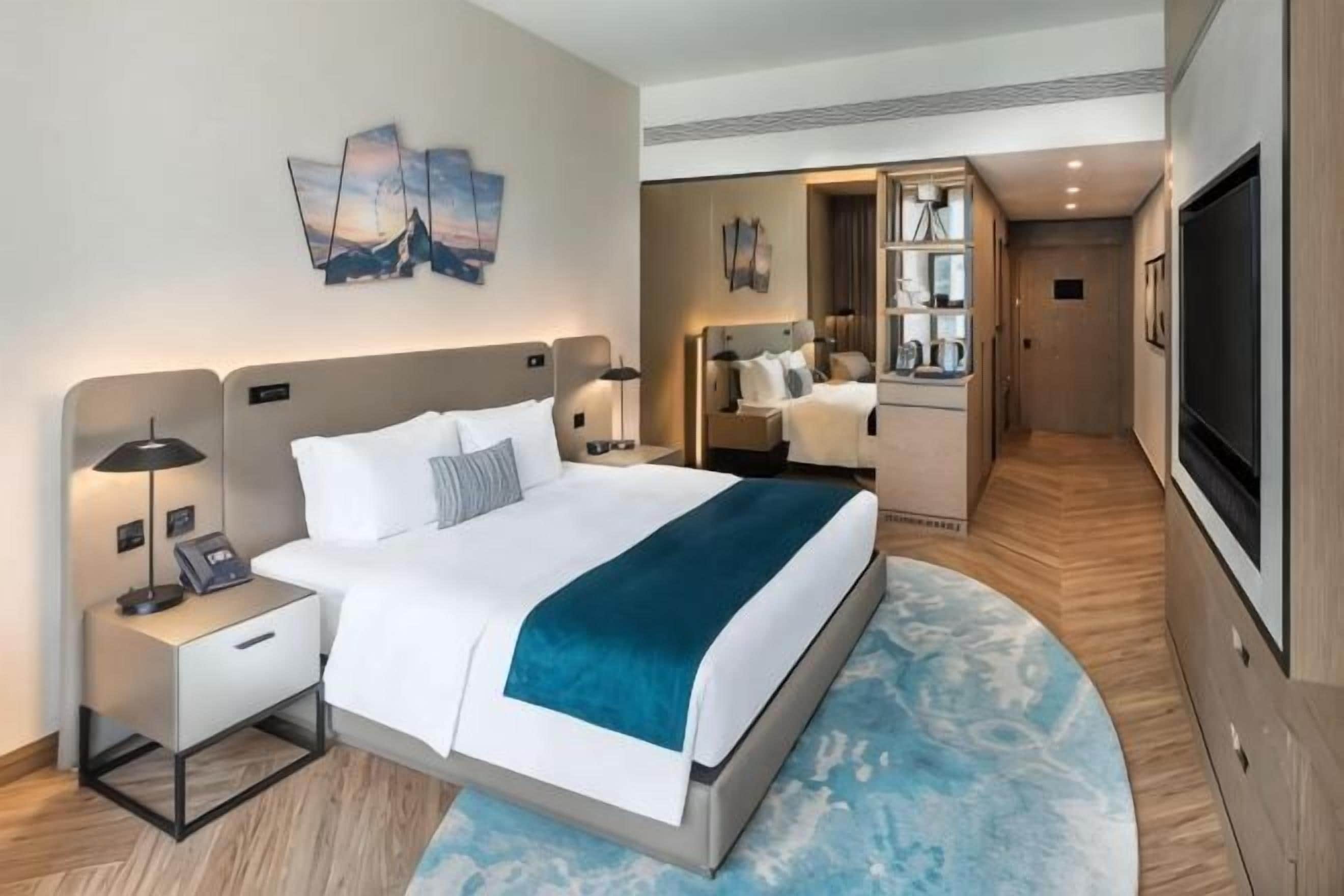A hotel room with a bed, bedside tables, a TV, a mirror, and a bathroom visible through an open door.