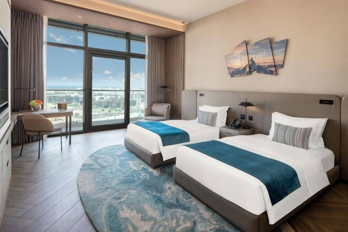 A hotel room with two beds, a desk, a chair, a large window, and artwork on the wall.