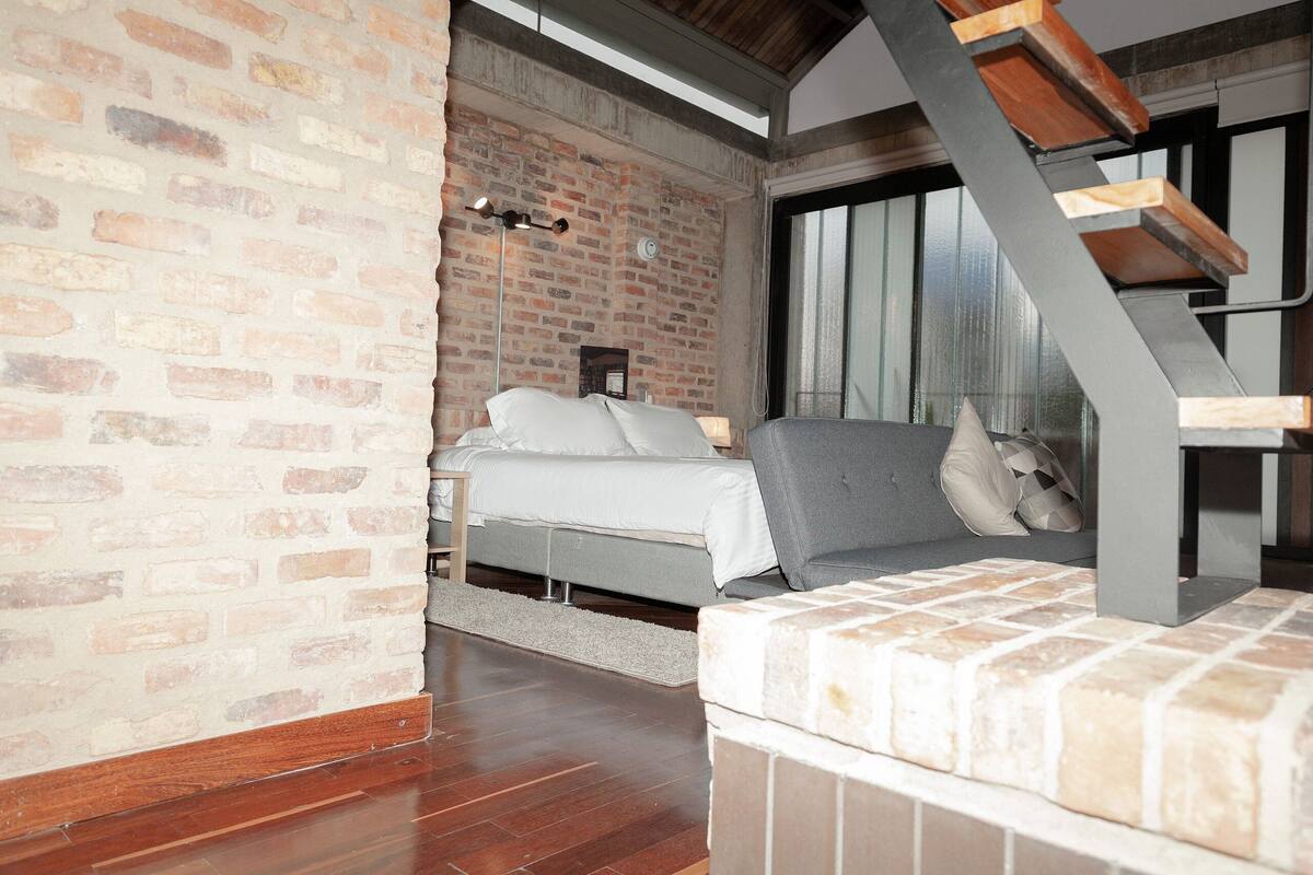 A bedroom with a brick wall, a bed, a grey sofa, and a wooden staircase.