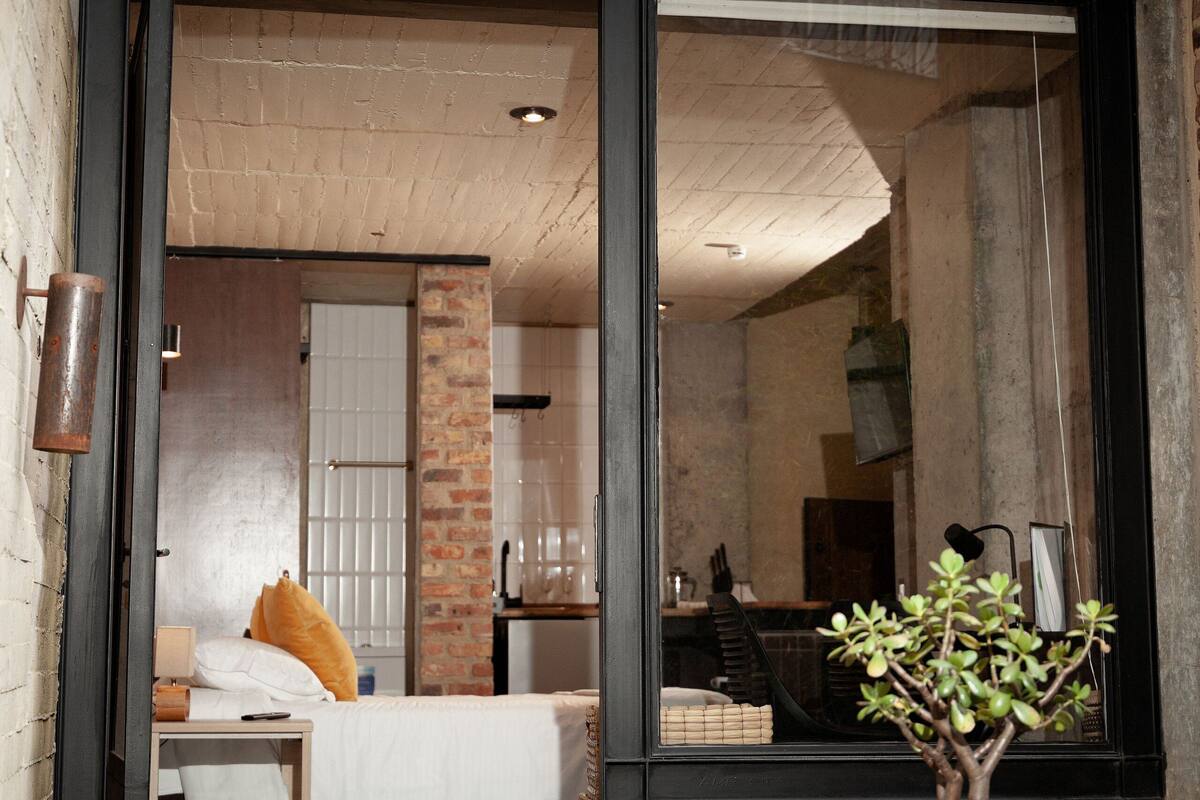 A modern room with a bed, a small table, and a window with a view of a brick wall and a staircase.