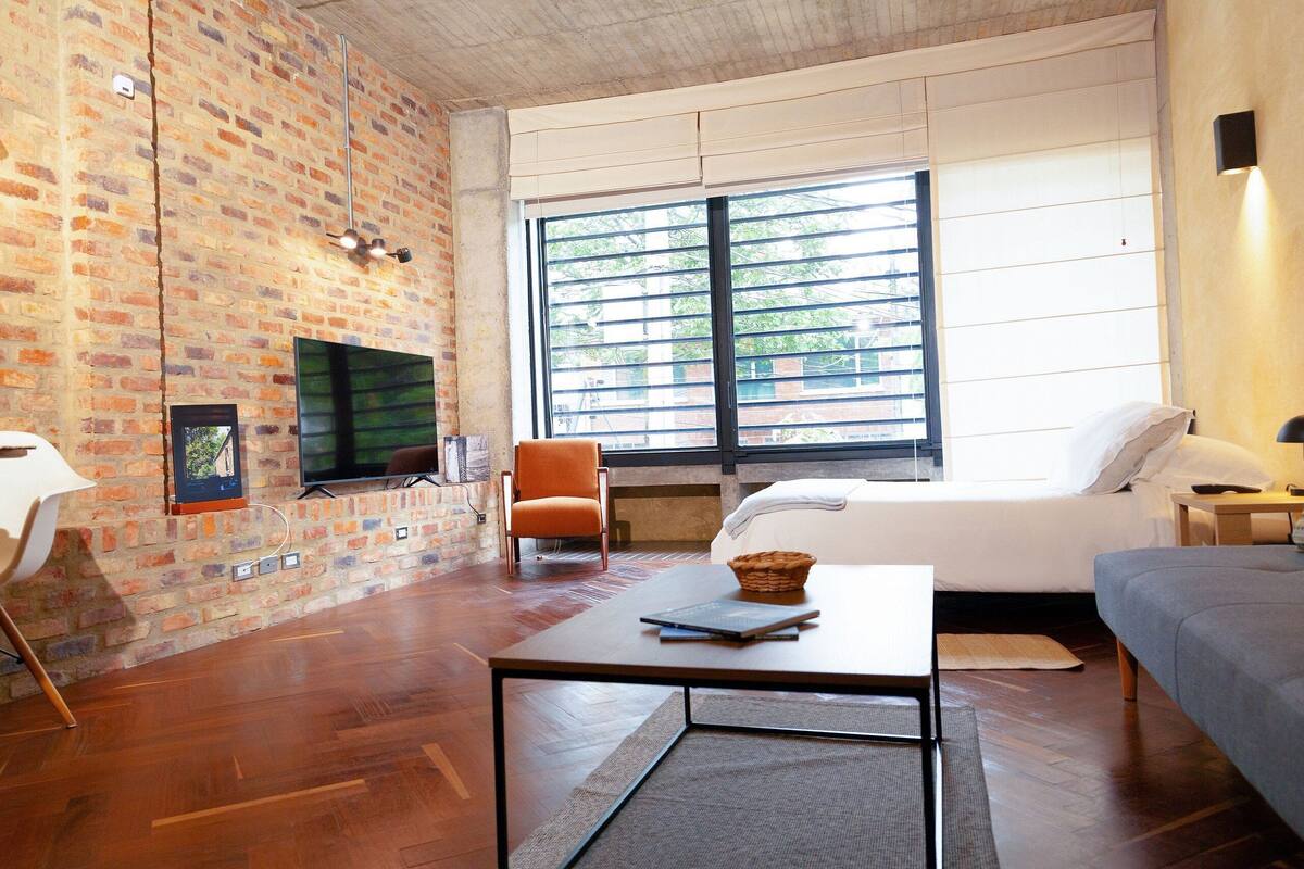 A modern living room with a brick wall, a large window, a sofa, a table, and a TV.