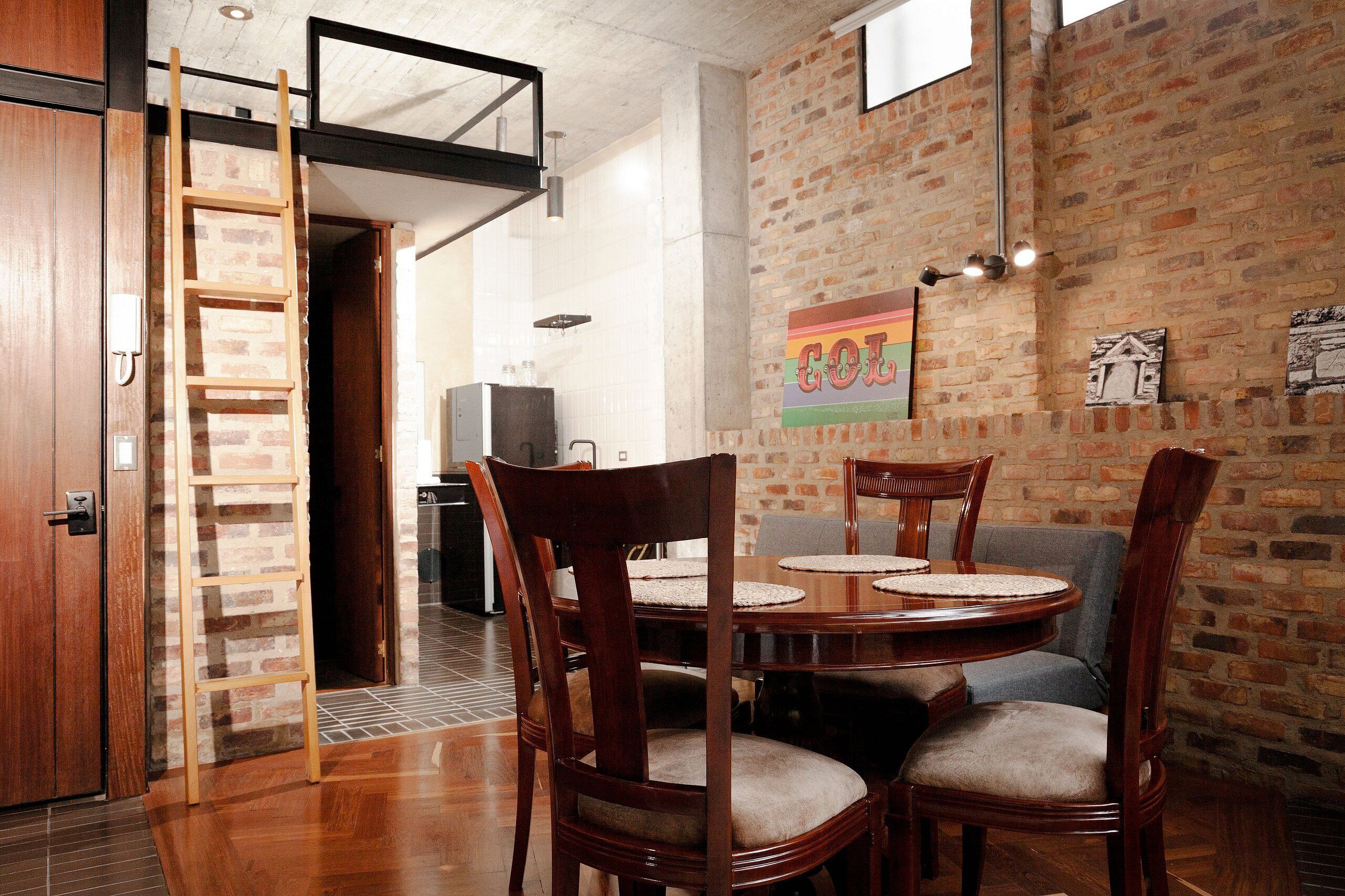 A dining area with a brick wall, wooden chairs, a round table, and a ladder against a door.