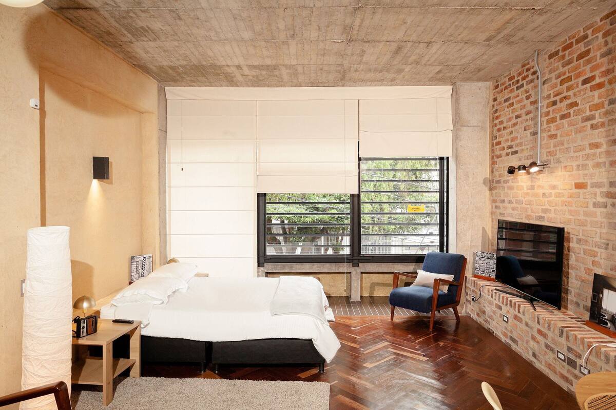 A modern bedroom with a large bed, a seating area with a fireplace, and a view of greenery through a window.