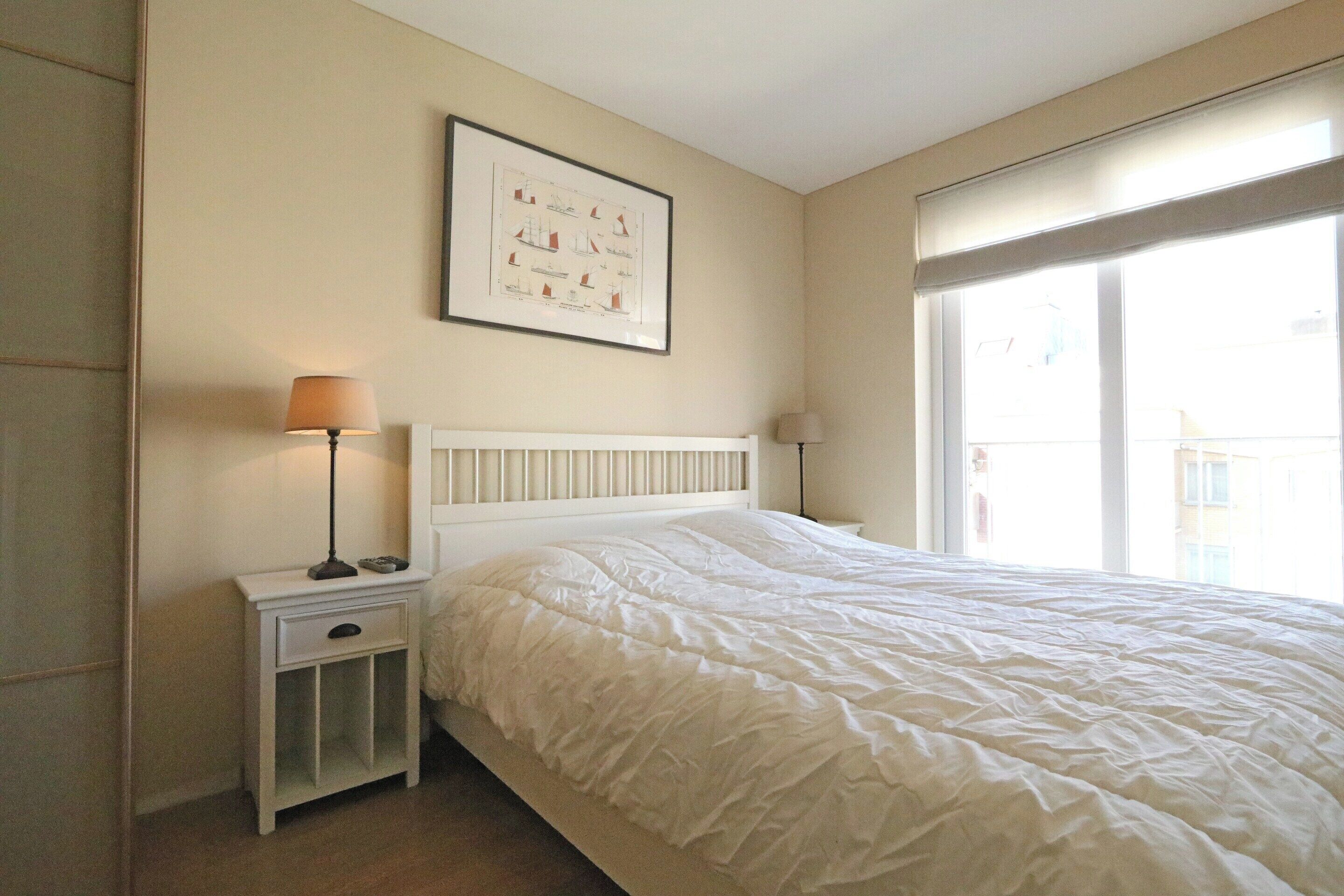 Une chambre avec un lit blanc, une table de chevet avec une lampe, un tableau encadré au mur et une fenêtre donnant sur un paysage.
