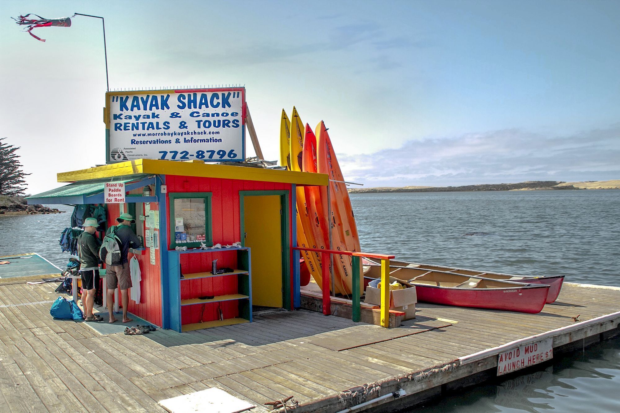 A colorful kayak shack with a signboard, paddleboards for rent, and a red kayak docked at a pier.