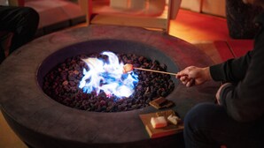 Une personne fait griller des guimauves au-dessus d’un feu de charbon de bois dans un espace extérieur aménagé en cercle avec des sièges.