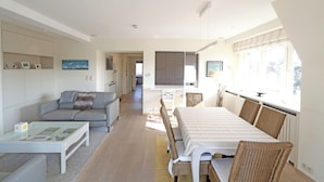 A modern living room with a sofa, coffee table, dining table, and chairs. There is a TV mounted on the wall and a window providing natural light. - Apartment in Knokke Near Zeedijk Beach (Knokke-Heist)