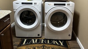 Un cuarto de lavandería con dos lavadoras y secadoras de carga frontal, gabinetes de madera en la parte superior y un tapete con la palabra 'Laundry'.
