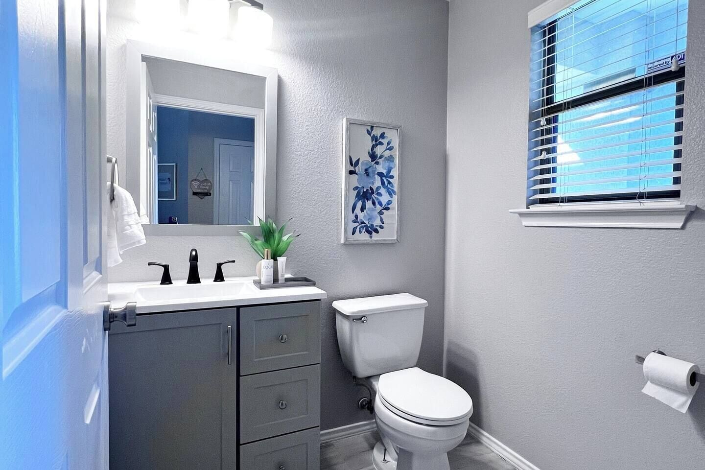 Un baño con inodoro blanco, lavamanos con grifos negros, un espejo sobre el lavamanos y una obra de arte floral enmarcada en la pared.