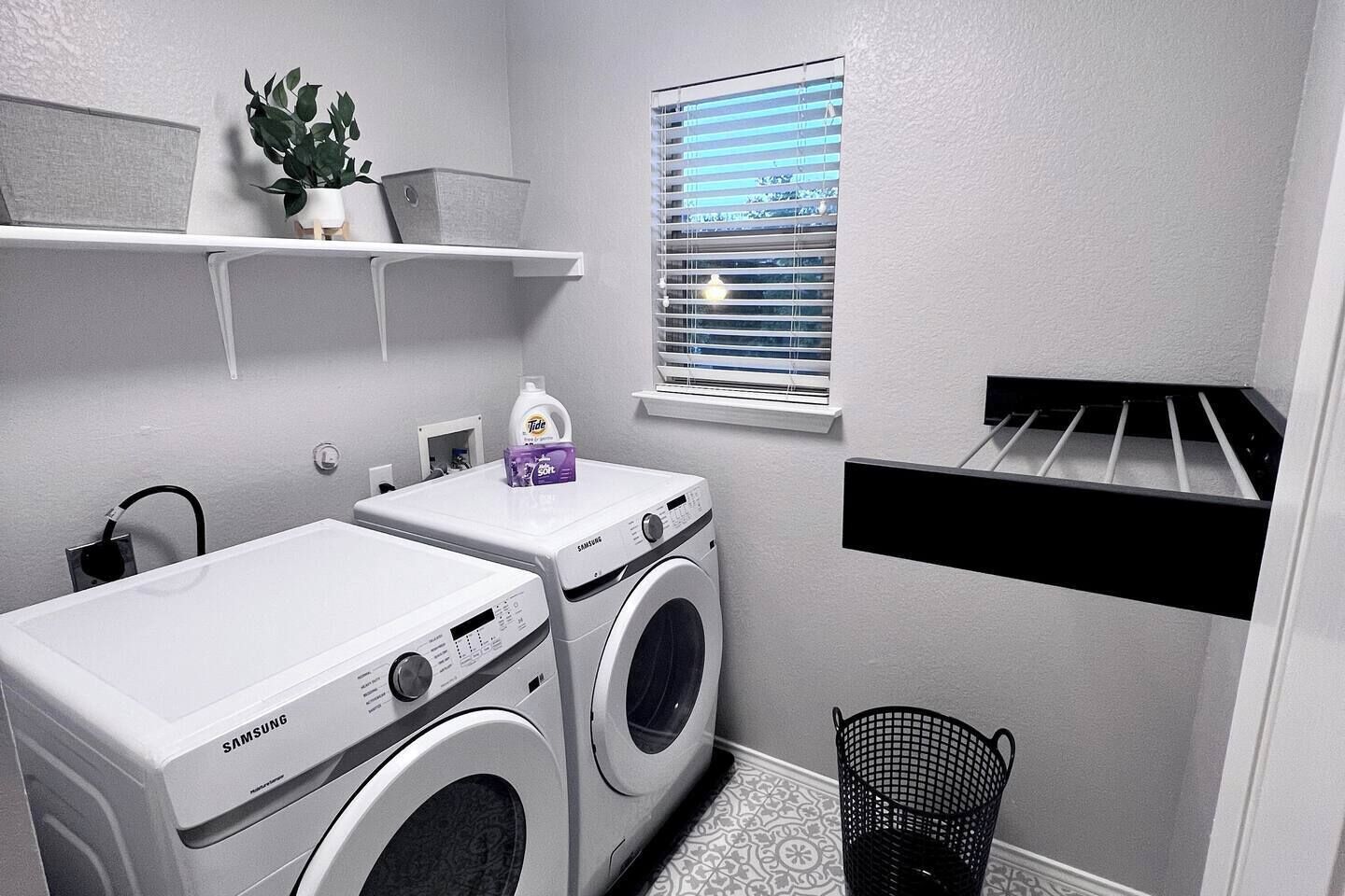 A compact laundry room with two Samsung washing machines, a window with blinds, a shelf with a plant, and a drying rack.