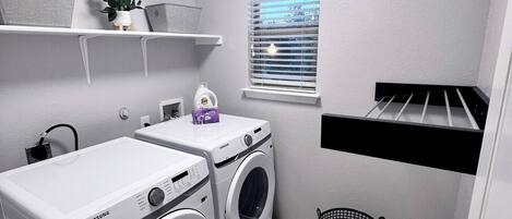 A compact laundry room with two Samsung washing machines, a window with blinds, a shelf with a plant, and a drying rack.