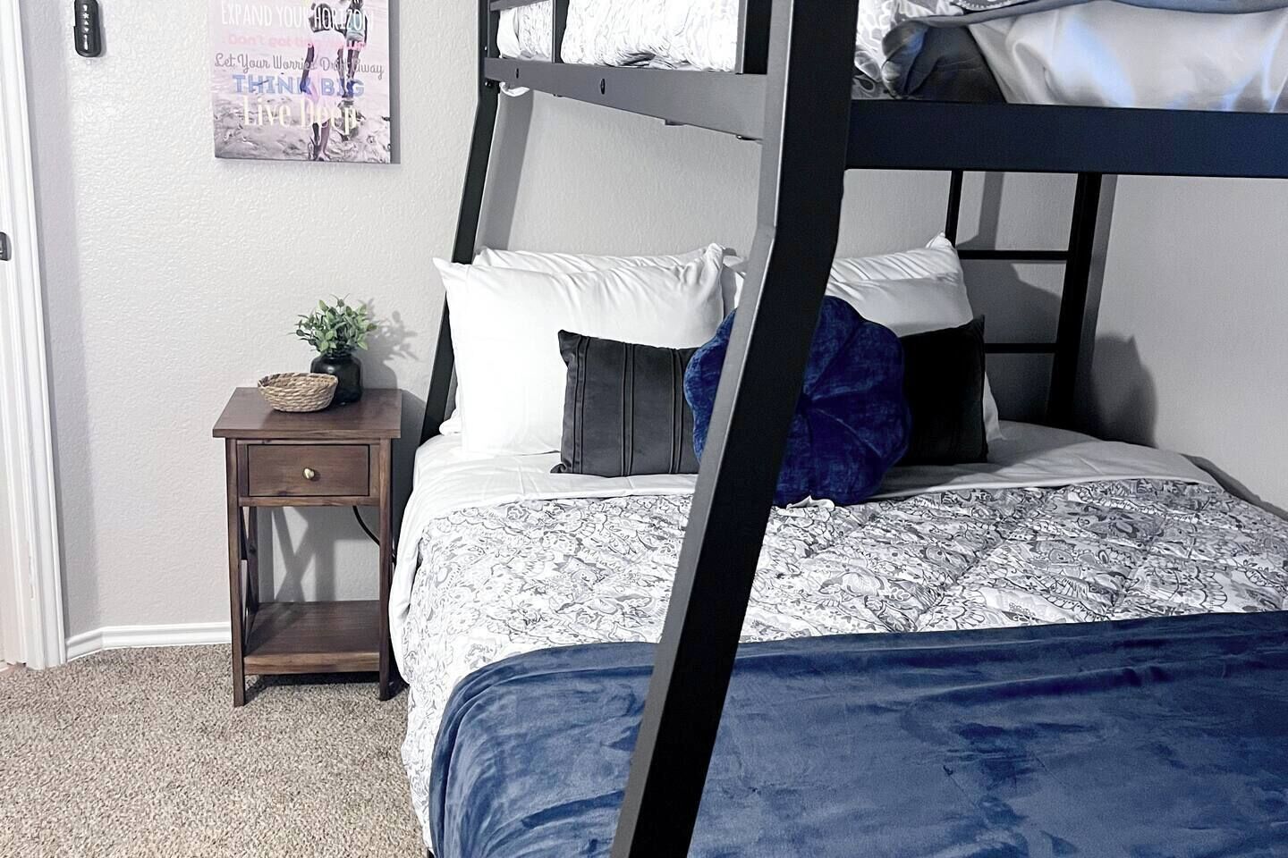 Ein Etagenbett mit gemustertem Bettbezug, ein Nachttisch mit Pflanze und ein motivierendes Bild an der Wand.