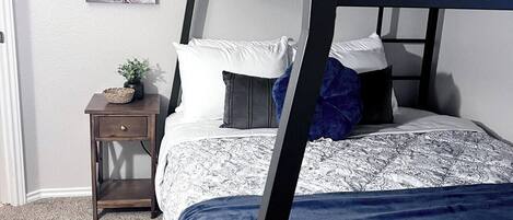 Ein Etagenbett mit gemustertem Bettbezug, ein Nachttisch mit Pflanze und ein motivierendes Bild an der Wand.