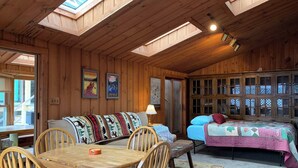 L'interno accogliente di una baita con un tavolo da pranzo in legno, sedie, un divano, un letto con copriletto e un lucernario.