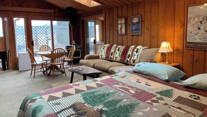 Un accogliente interno di una baita con soffitto in legno, un divano, una coperta trapuntata e una zona pranzo con sedie e tavolo.