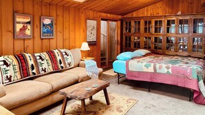 Un accogliente interno di una baita con soffitto in legno, un divano con coperte decorate, un letto con copriletto e un armadio in legno con ante in vetro.