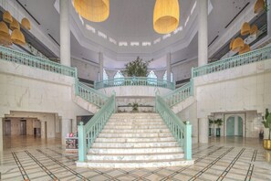 Eine imposante Treppe mit Marmorstufen und türkisblauen Geländern, die zu einem Balkon im zweiten Obergeschoss führt.