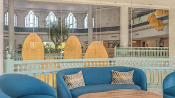 Un hall d’entrée spacieux, agrémenté d’un canapé bleu, de lampes en rotin suspendues et offrant une vue sur un bâtiment traditionnel aux toits de tuiles.