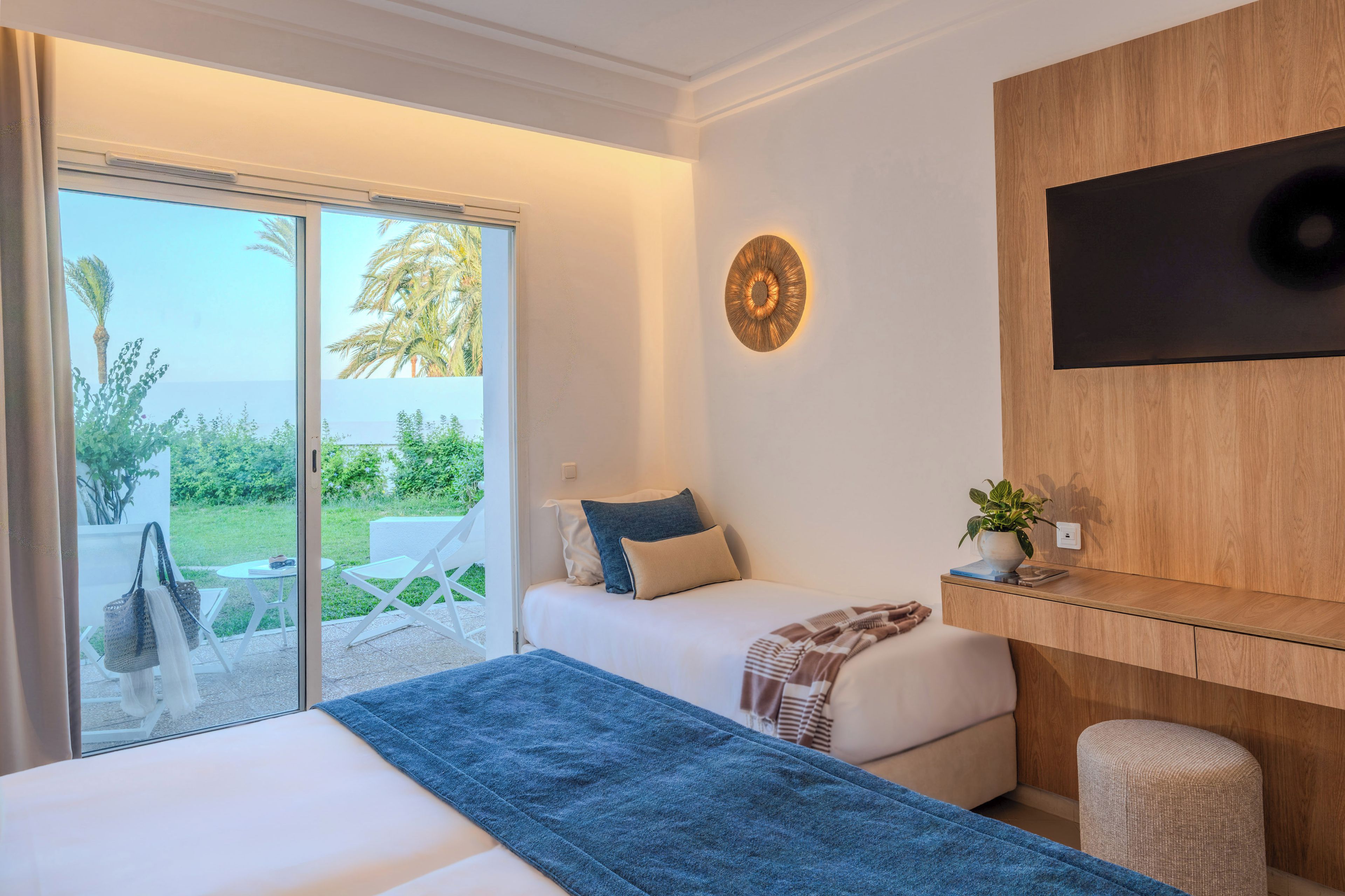 A hotel room with a bed, a TV mounted on the wall, a small table with a plant, and a view of the outdoors through a sliding door.