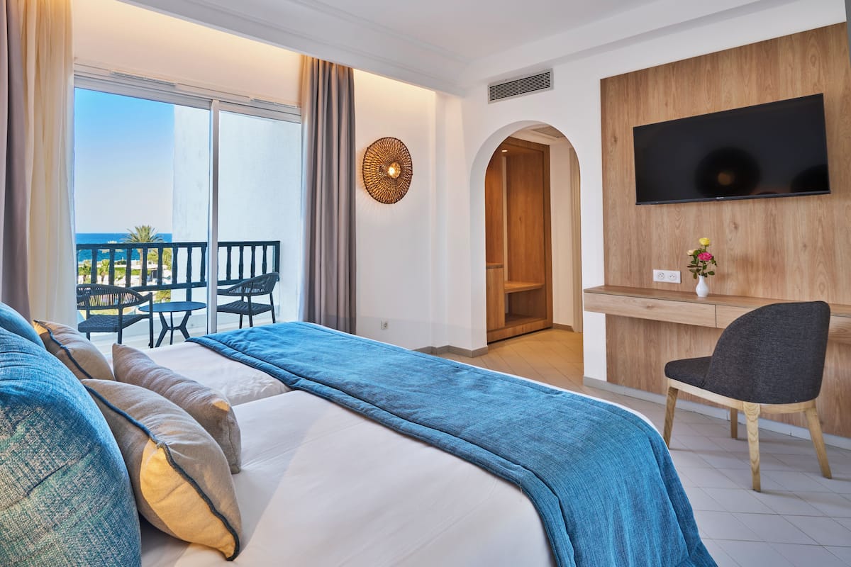 Habitación de hotel con cama, escritorio, silla, televisión y balcón con vista al mar.