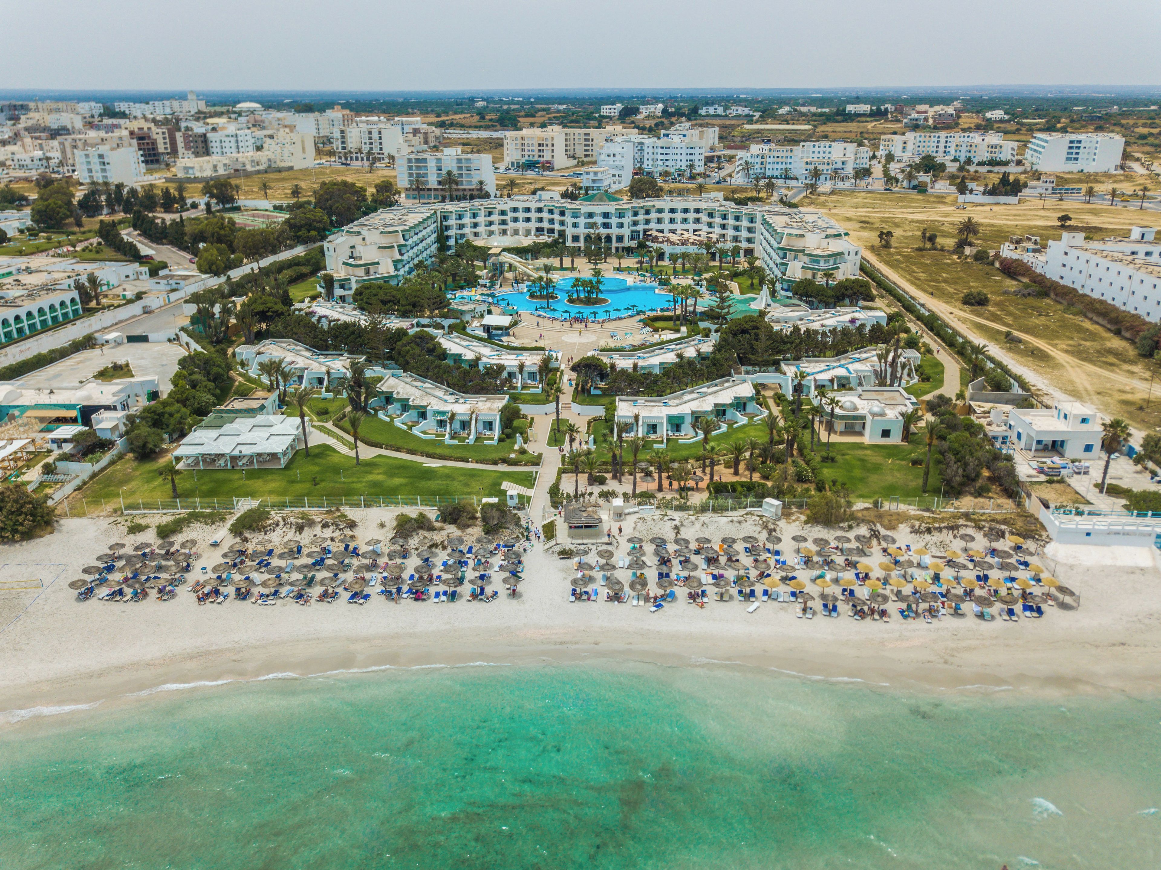 An aerial view of a resort with a central pool, multiple buildings, and a beach area with sun loungers.