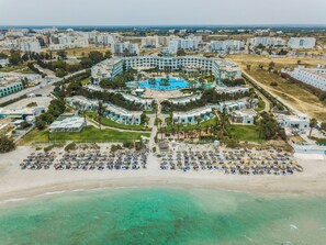 Eine Luftaufnahme eines Resorts mit einem zentralen Pool, mehreren Gebäuden und einem Strandbereich mit Liegestühlen.