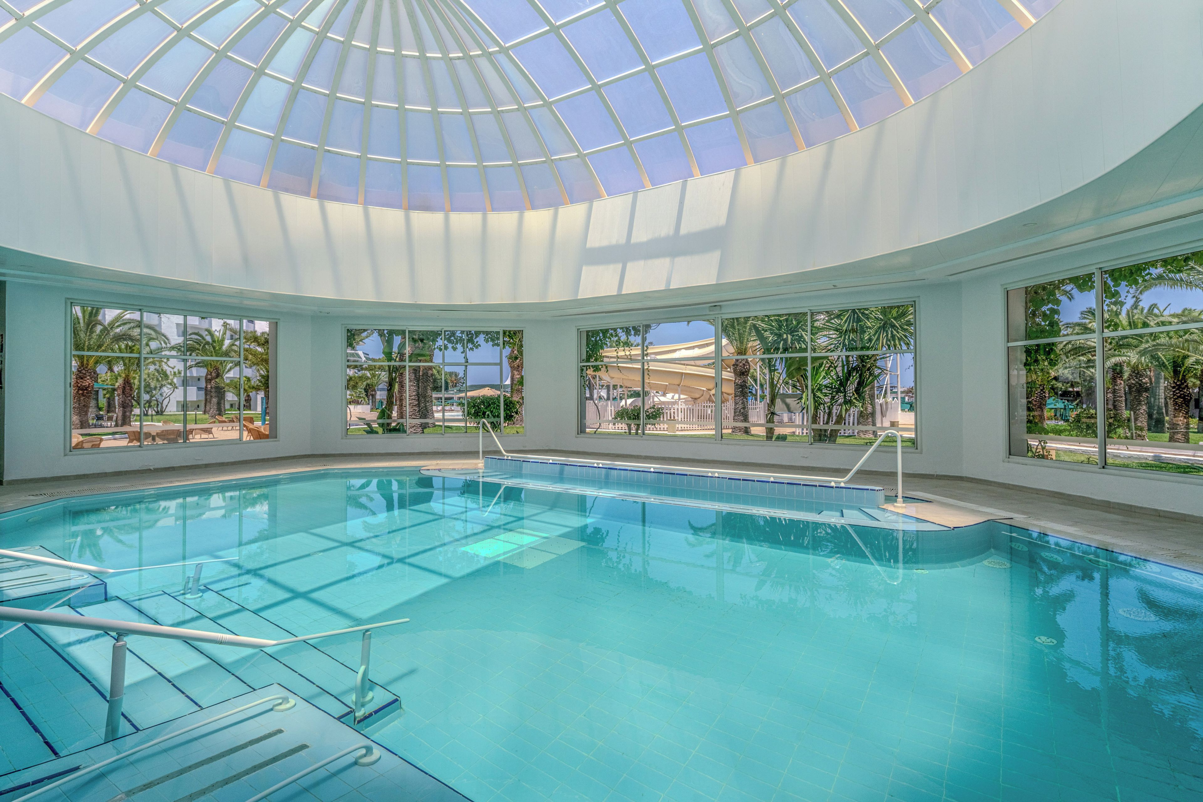 Una piscina interna con soffitto in vetro, acqua cristallina e vista su palme e edifici esterni.
