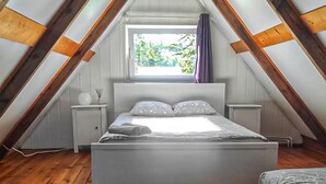 Een slaapkamer met een schuine plafond, een bed met kussens, een nachtkastje met een lamp en een raam met gordijnen.